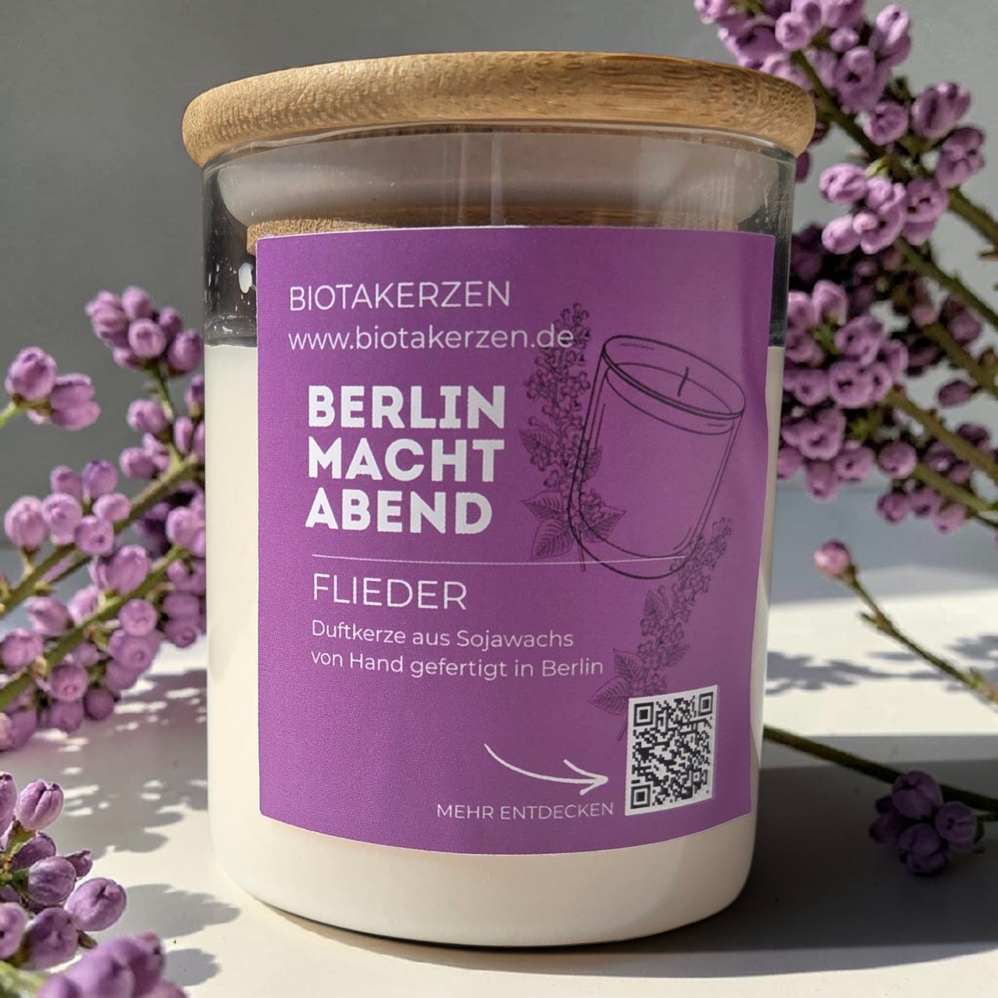Duftkerze Flieder von BiotaKerzen aus der Kollektion Berlin macht Abend, im Glas mit Bambusdeckel, dekoriert mit lila Blüten.